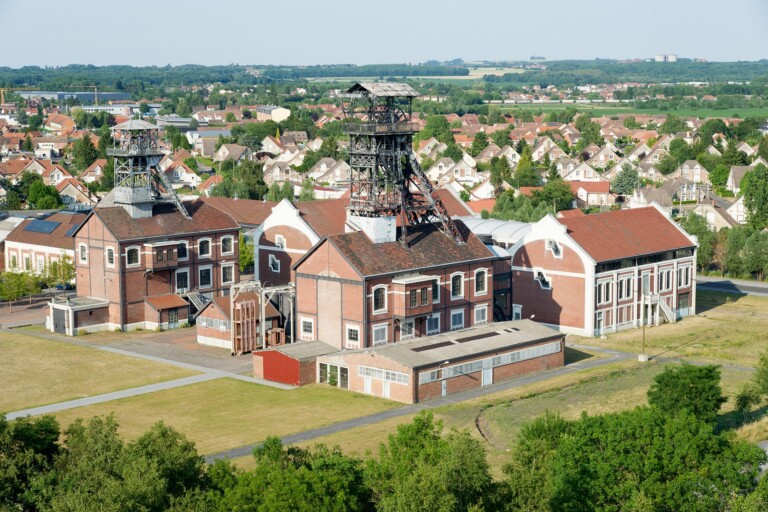 Complexe industriel historique avec bâtiments en briques rouges et tours de mine, entouré de verdure et de maisons résidentielles en arrière-plan