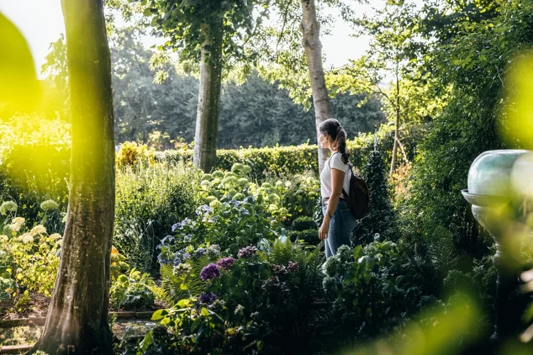 femme debout dans un jardin luxuriant entouré de plantes et d'arbres, avec la lumière du soleil filtrant à travers le feuillage