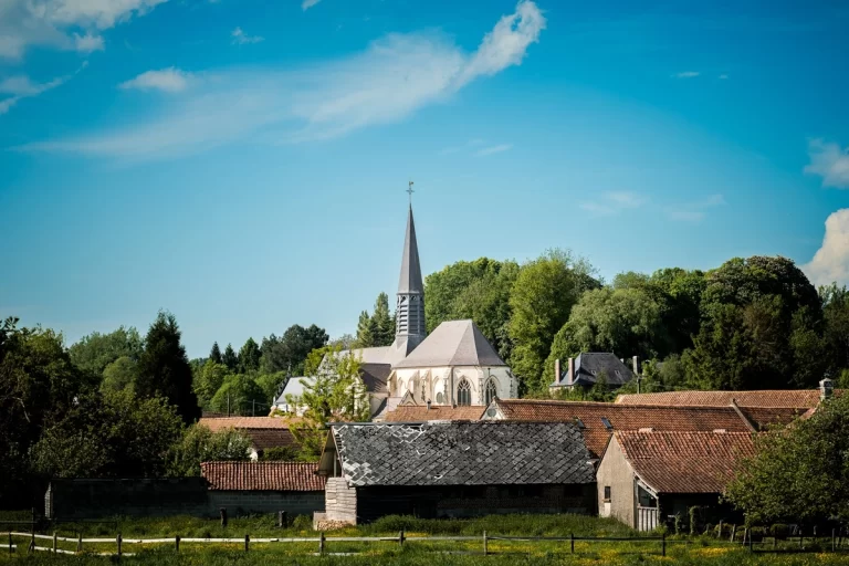 Village pittoresque avec une église au clocher pointu au centre, entourée de bâtiments aux toits en tuiles rouges et d'arbres verdoyants sous un ciel bleu partiellement nuageux